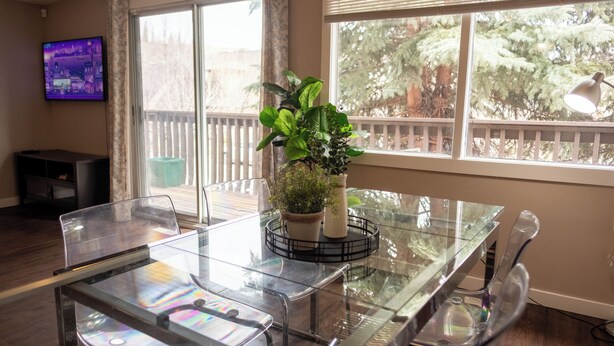 A modern dining area with a glass table, clear chairs, a potted plant, and a TV on the wall. - Near Riverside Sunset-Lodge w/Backyard+BathTub-W/D (Calgary)