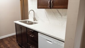 A kitchen with dark wooden cabinets, a white countertop, a stainless steel sink, and a white dishwasher. - Near Riverside Sunset-Lodge w/Backyard+BathTub-W/D (Calgary)