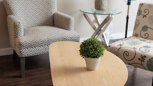 A modern living room with a patterned armchair, a glass coffee table with a potted plant, and a standing fan. - Near Riverside Sunset-Lodge w/Backyard+BathTub-W/D (Calgary)