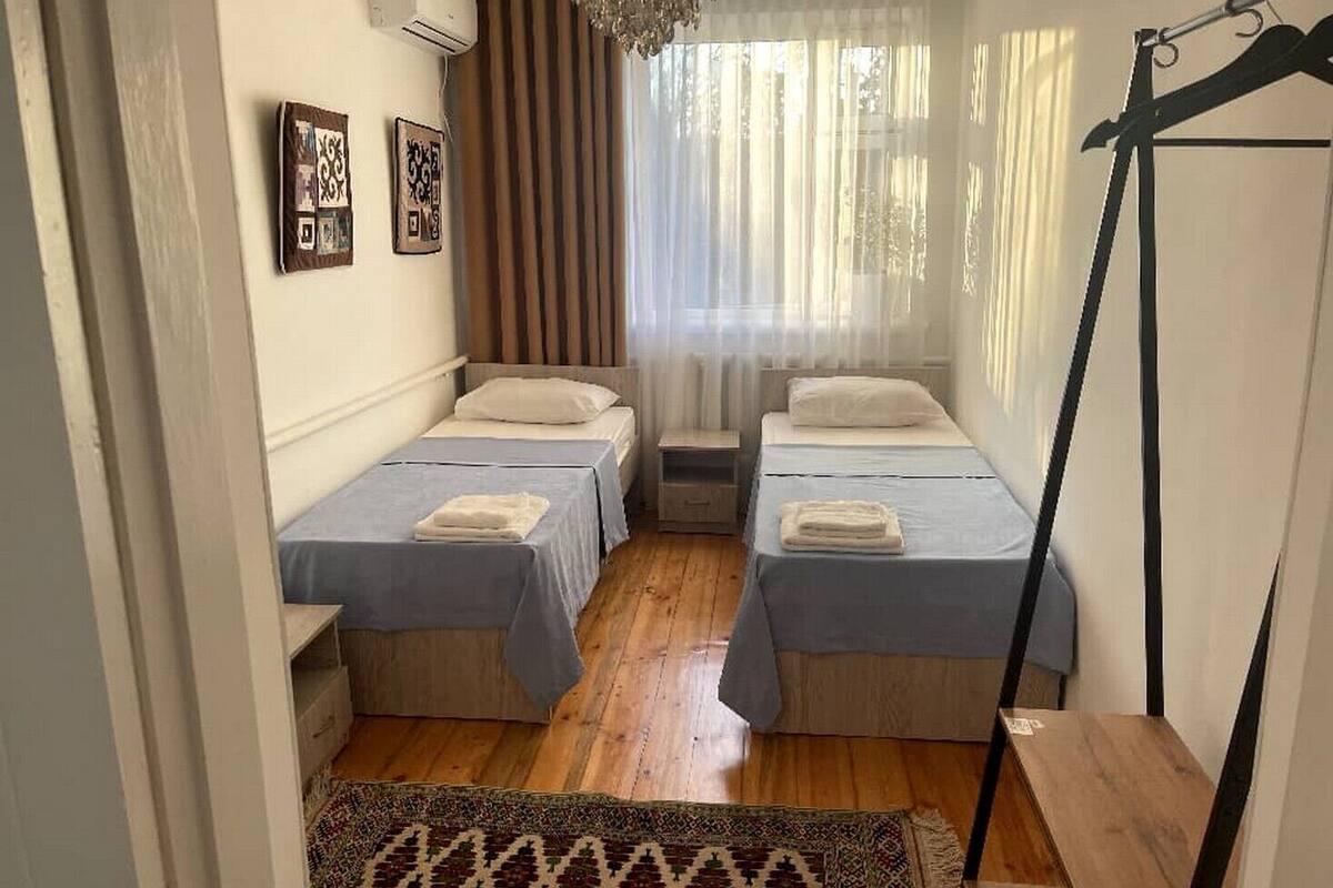 A small, cozy room with two single beds, a wooden floor, a patterned rug, and a window with sheer curtains.