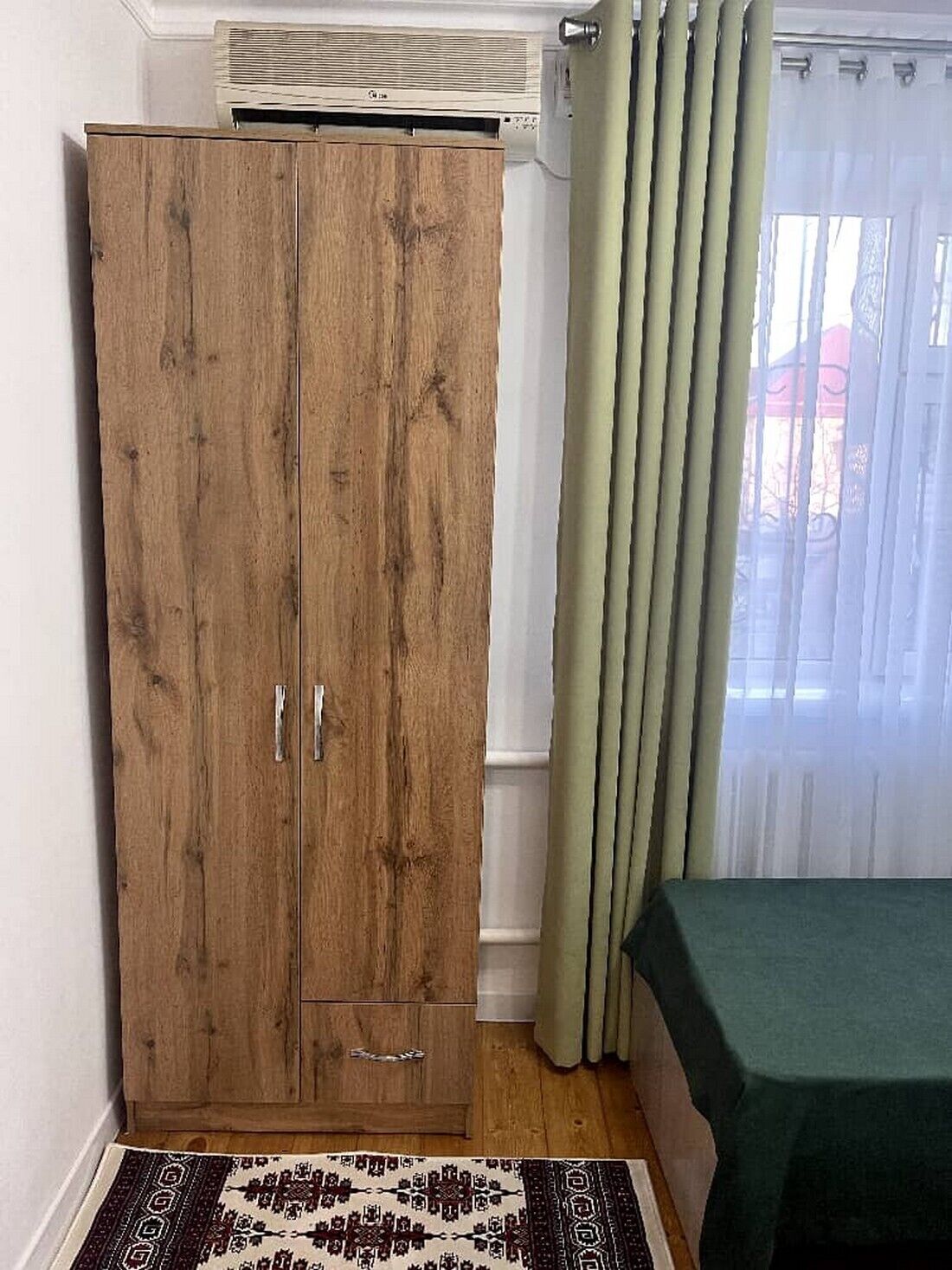 Een kamer met een houten garderobe, een bed met een groene spreidekking, een kamerbreed tapijt met een patroon en een raam met gordijnen.