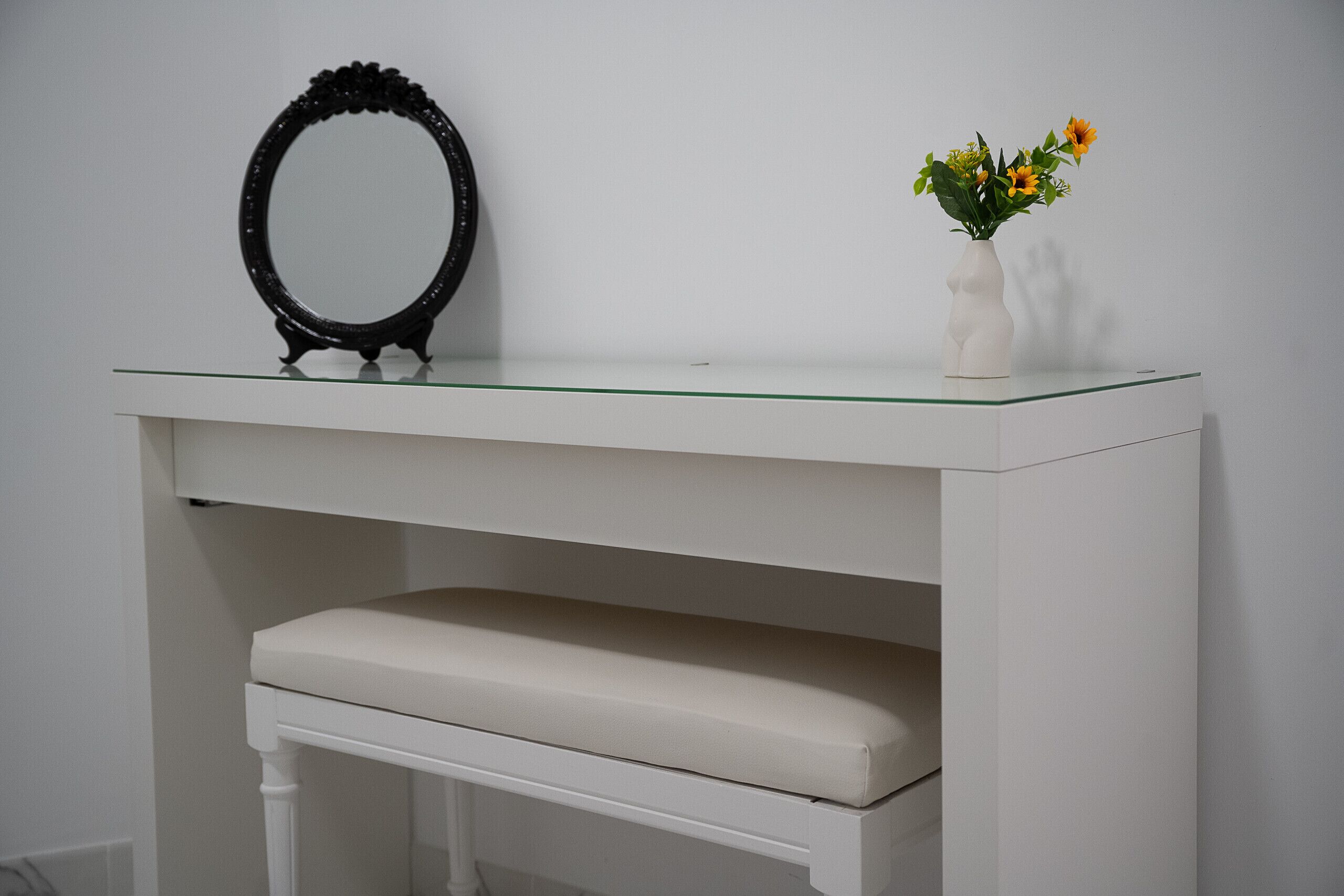 A white vanity with a glass top, a black ornate mirror, and a white vase with flowers.