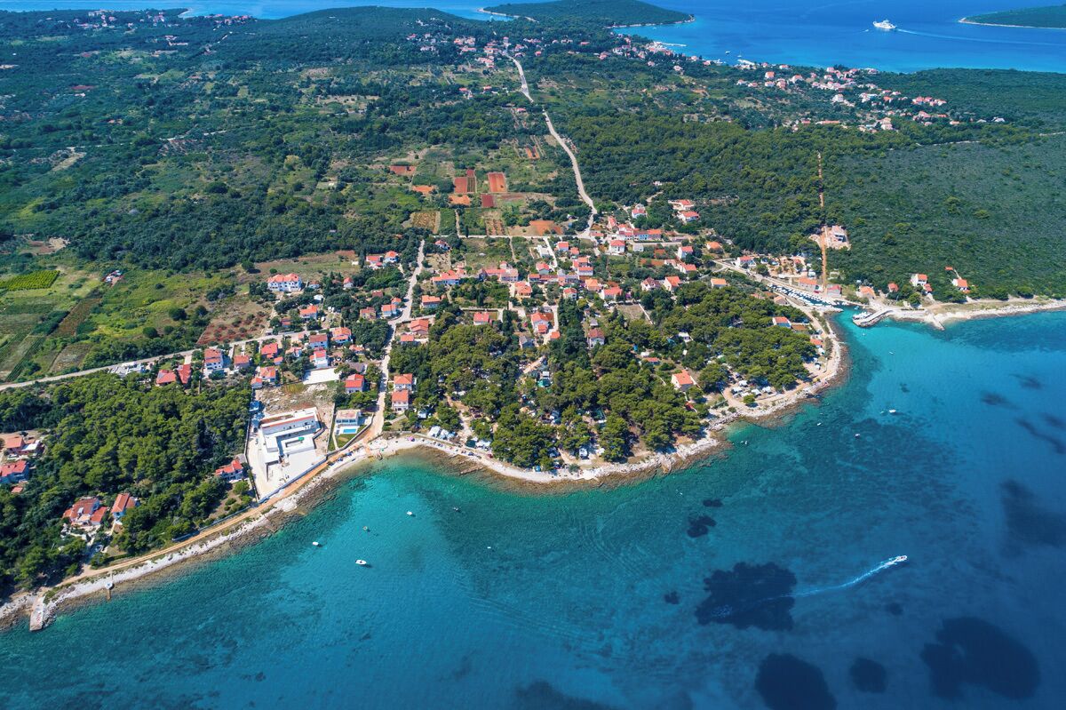 An aerial view of a coastal town with a dense cluster of red-roofed buildings, a prominent white building with a blue roof, and a curved coastline with clear blue waters.