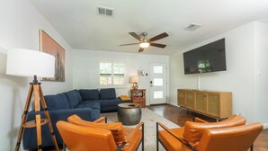 A living room with a blue sofa, orange chairs, a wooden cabinet, and a ceiling fan. - Stylish Group Retreat • Hot Tub • Game Room • Yard (Austin)