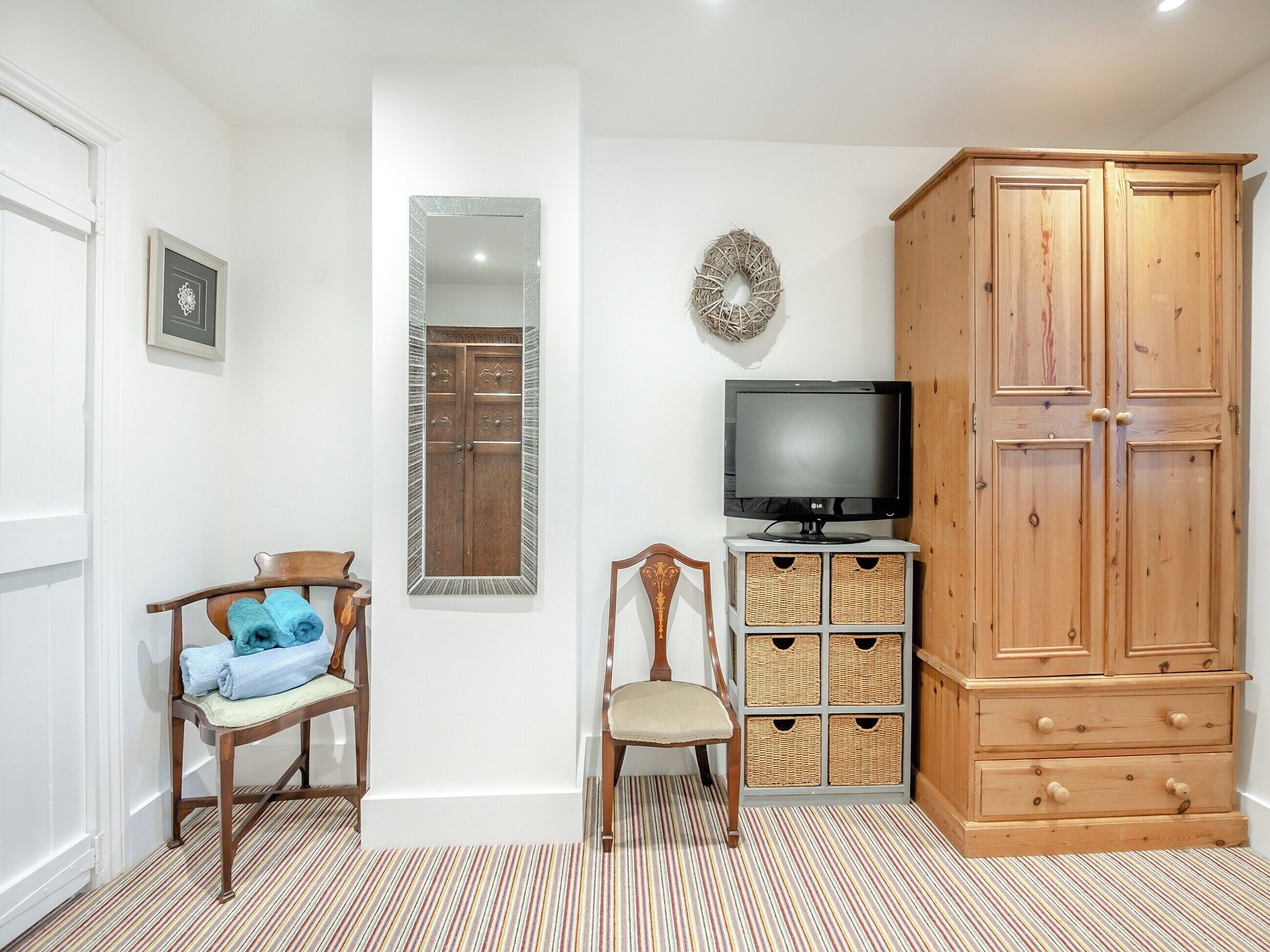 A room with a wooden wardrobe, a television on a stand, a chair with a cushion, a small table, and a mirror on the wall.