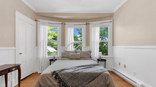A bedroom with a large bed, two nightstands, a wooden table, and three windows with white curtains. - Studio 5~ Cozy BDRM w/ Private Bath (Moncton)