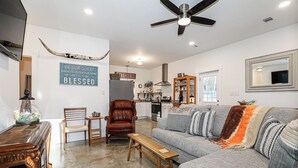 Ein geräumiges Wohnzimmer mit grauem Sofa, Couchtisch aus Holz und Ledersessel. Im Hintergrund ist eine Küche erkennbar, mit einem Schild, auf dem steht: "BE OUR GUEST WHEN YOU ENTER OUR HOME MAY YOU BE BLESSED.".