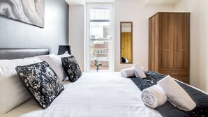 A hotel room with a bed, pillows, a wooden wardrobe, and a window with a view of buildings. - From Home to Home (Birmingham)