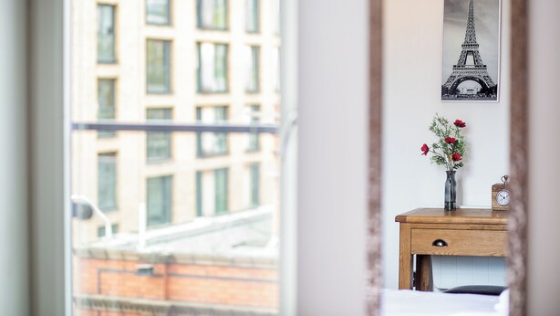 A room with a view of a building through a window, a wooden bedside table with a vase of flowers, and a framed picture of the Eiffel Tower on the wall. - From Home to Home (Birmingham)