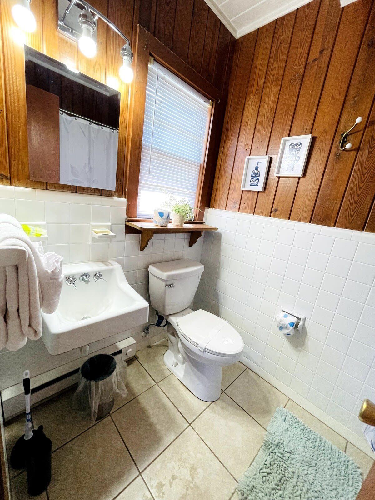 Ein Badezimmer mit Holzpaneelen, einem weißen Waschbecken, einer Toilette und einem Fenster mit Jalousien.