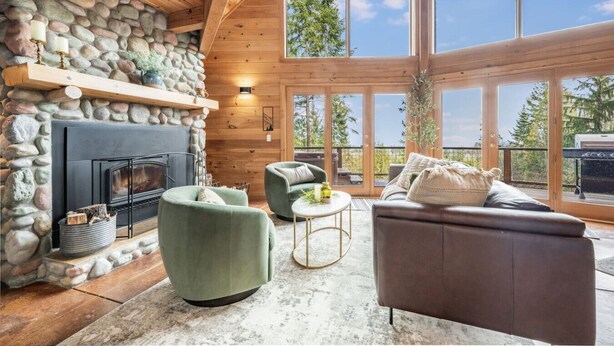 Interieur van een blokhut met een stenen open haard, groene fauteuils, een bruine bank en grote ramen met uitzicht op bomen.