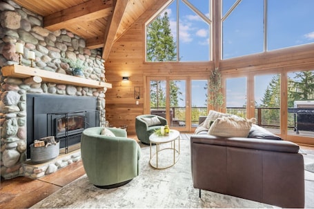 A log cabin interior with a stone fireplace, green armchairs, a brown sofa, and large windows offering a view of trees.