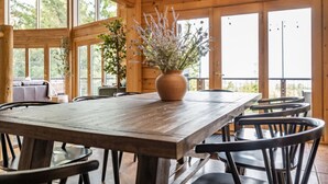 Een houten interieur met een grote tafel en stoelen, een vaas met bloemen en grote ramen.