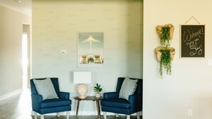 A room with two blue armchairs, a small table with a lamp, a framed picture of a beach scene, and a welcome sign with greenery. - Charming Brand New Home in Foley (Foley)