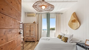 Un dormitorio con un armario de madera, una cama con ropa de cama blanca, un sombrero original sujeto a la pared y vistas a través de una ventana.