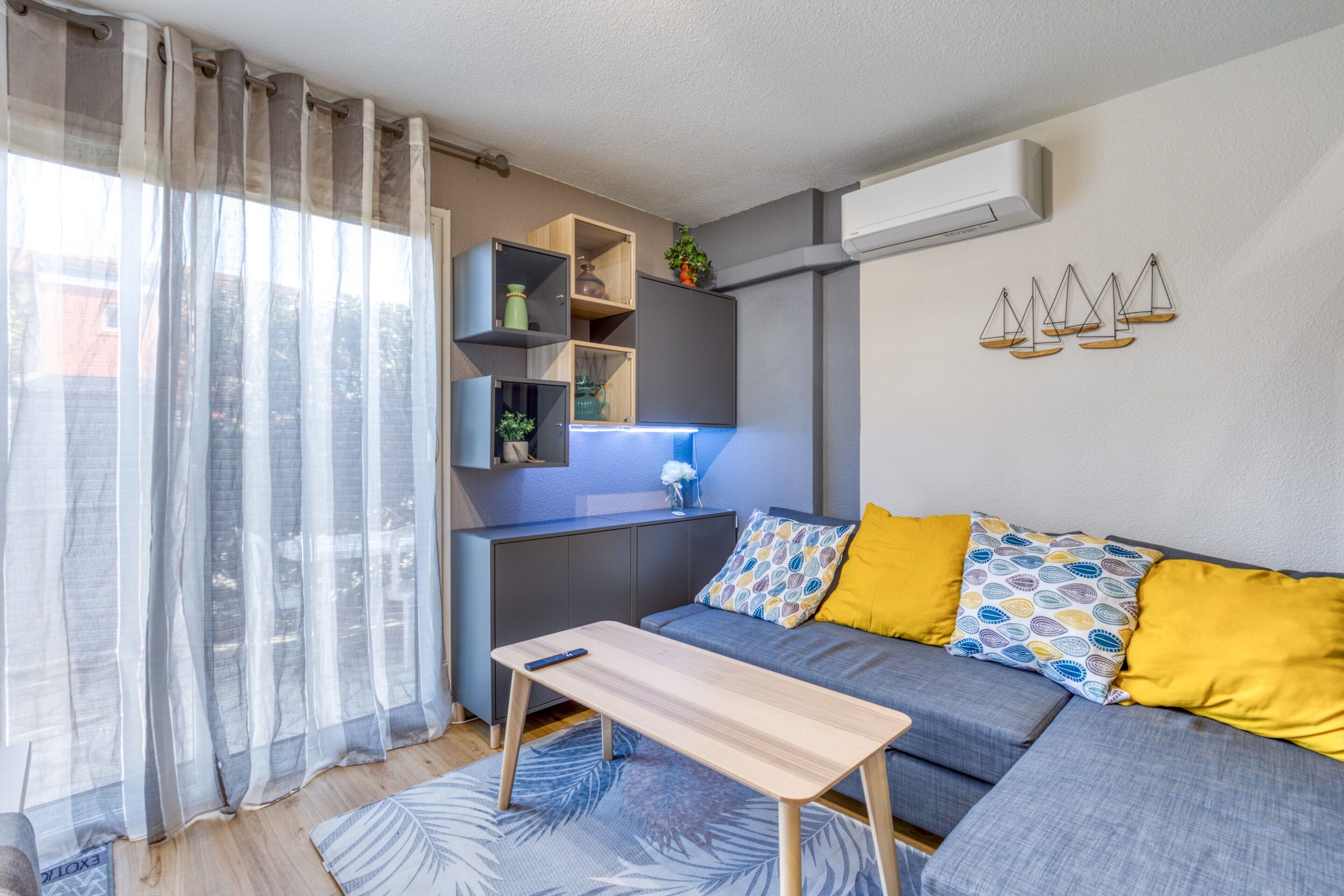 Una sala de estar moderna con un sofá, una mesita y una zona de cocina con estantes y aire acondicionado.