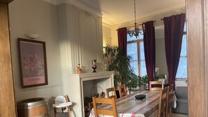 Ein Esszimmer mit Kamin, Holzsesseln, einem Tisch mit kariertem Tischläufer und einem großen Fenster mit Vorhängen.