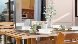 Une table de salle à manger en bois, dressée avec de l’assiette blanche, des bols et un vase contenant des feuilles vertes.