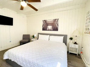 Un dormitorio con una cama grande, un ventilador de techo, un televisor montado en la pared y una mesita de noche con una lámpara y una planta pequeña.