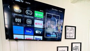 A flat-screen TV mounted on a white wall displaying various streaming services. Below the TV, there is a console table with decorative items and framed motivational quotes. - Cozy one bedroom across from the beach #2 (North Myrtle Beach)