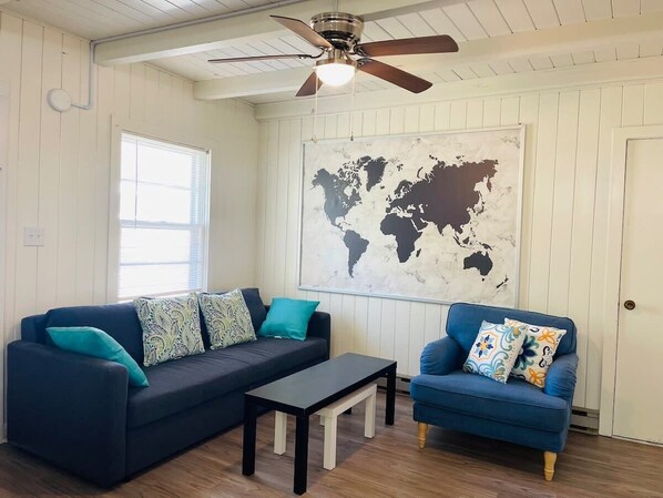 Una habitación con un sofá azul oscuro, un sillón a juego y una mesita pequeña. Un mural de un mapa mundial adorna la pared, y hay un ventilador de techo.