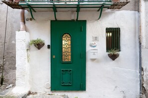 Una puerta verde con un panel de vidrio decorativo, una pequeña ventana con una reja de metal y dos macetas con plantas a cada lado.