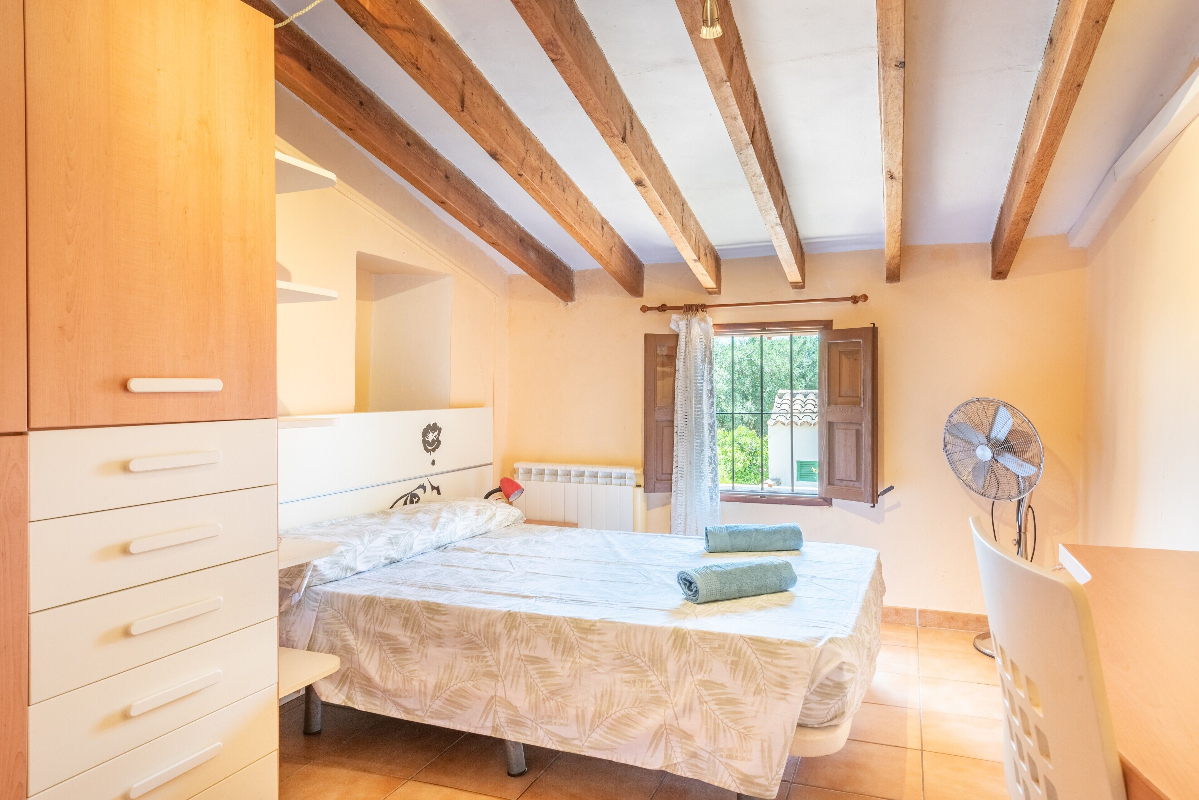 Ein Schlafzimmer mit Bett, Holzbalken, Heizkörper, Fenster mit Vorhängen und einem Standventilator.