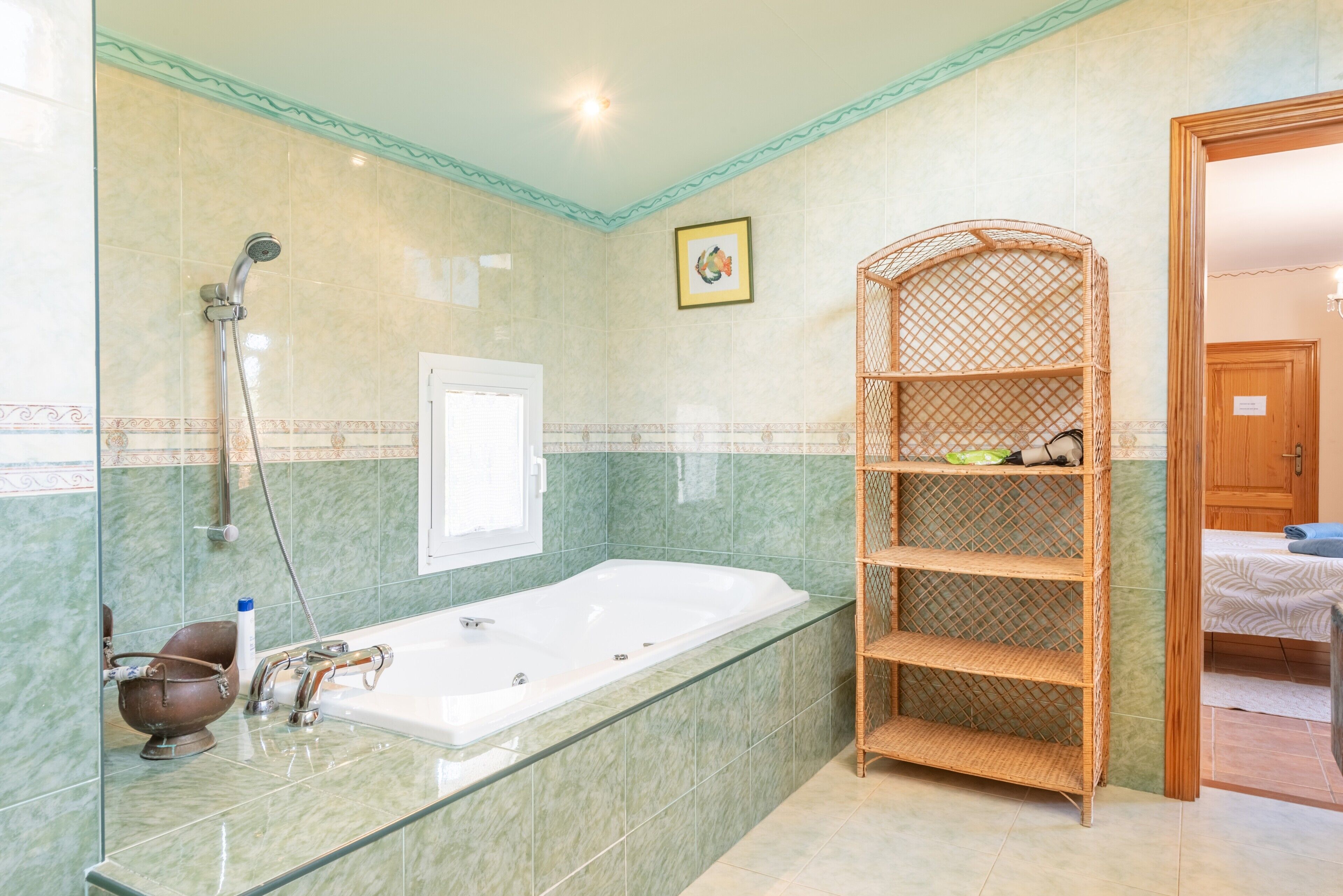 A bathroom with a glass-enclosed bathtub, a shower, a wicker shelf, and a door leading to another room.