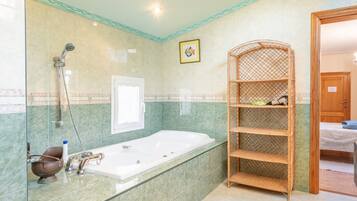 A bathroom with a glass-enclosed bathtub, a shower, a wicker shelf, and a door leading to another room.