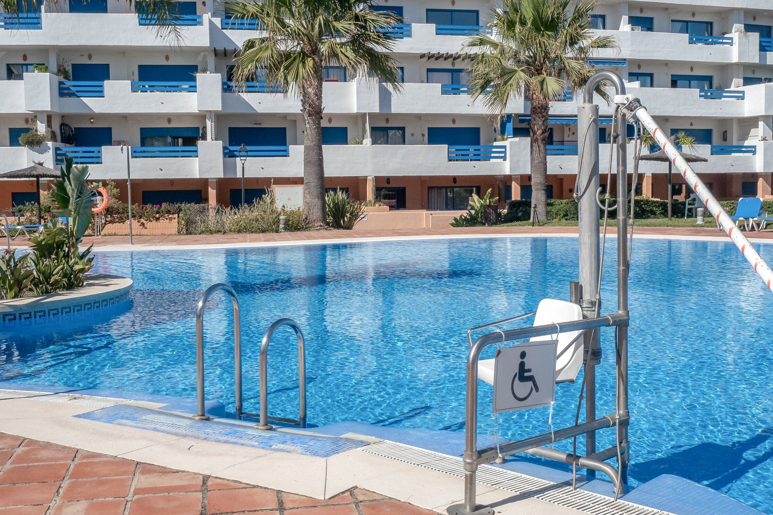 A swimming pool with a lifeguard stand, a wheelchair accessibility sign, and a building with balconies in the background.