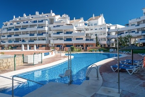 Une piscine dotée d’un parasol, de chaises longues et d’un bâtiment avec des balcons.
