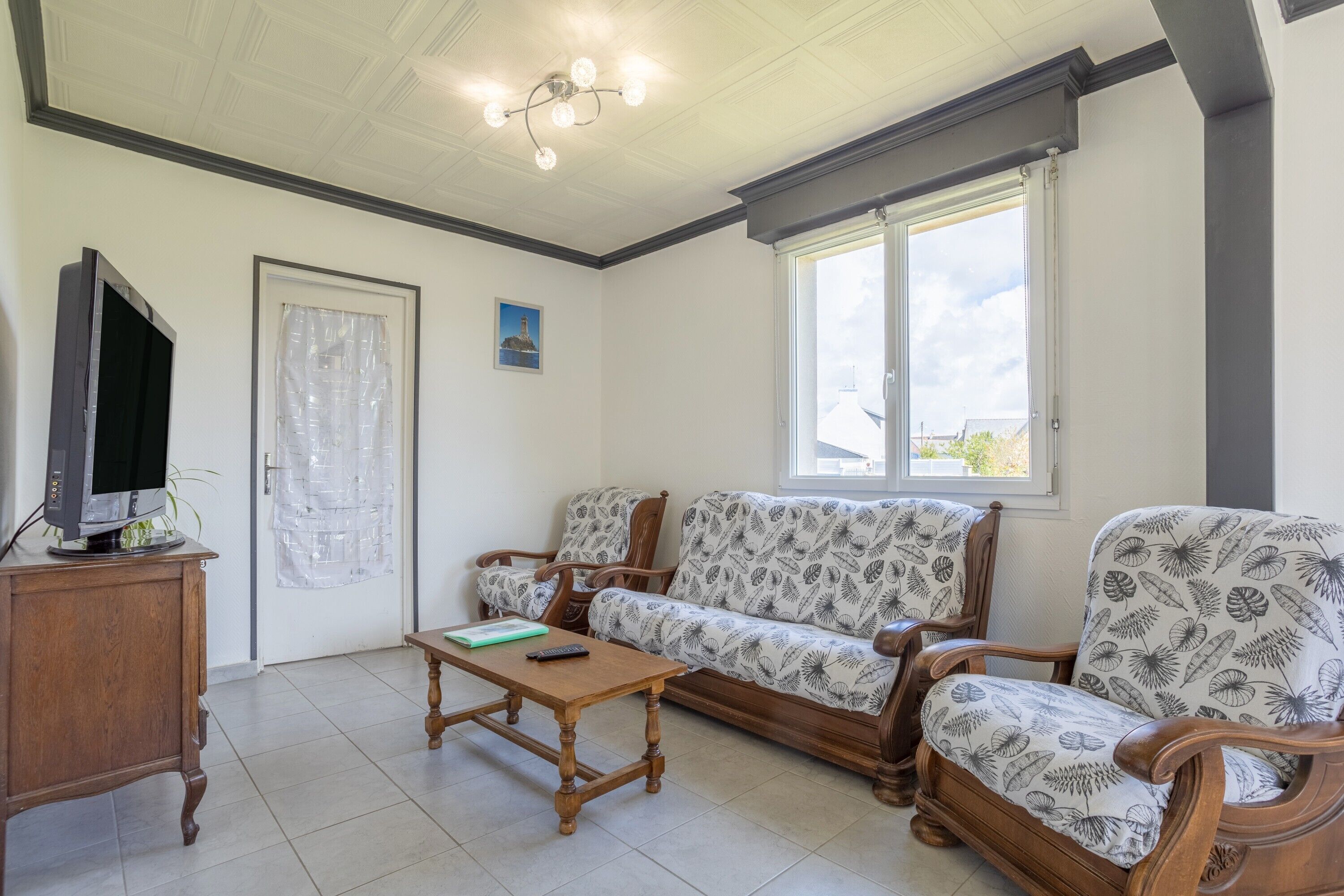 Un salon comprenant un canapé, une table basse en bois, un téléviseur posé sur un meuble, une porte avec un rideau et une fenêtre donnant sur un paysage agréable.