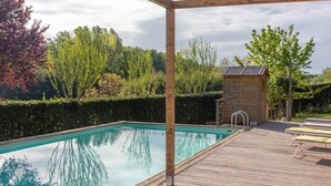 En swimmingpool med en træterrasse, en mindre trækonstruktion og en hæk i baggrunden.