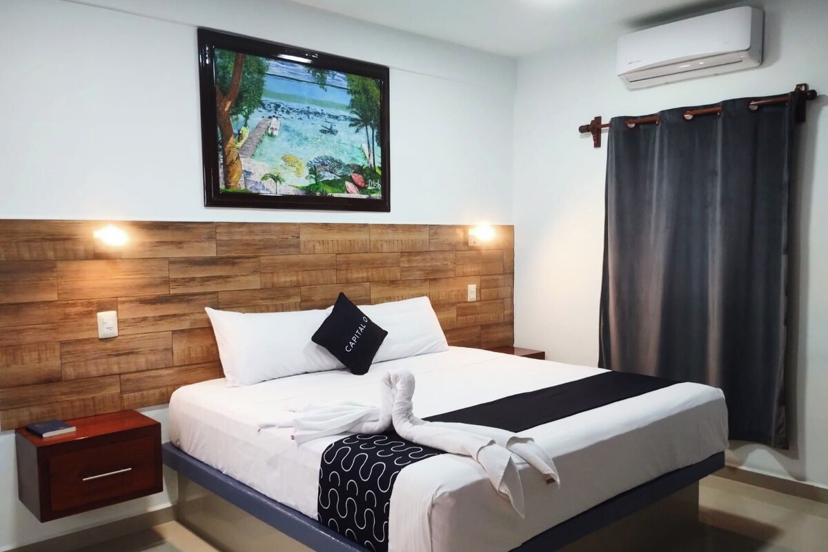 A hotel room with a wooden headboard, a bed with white bedding, a black pillow, a black and white patterned throw, a white towel, a small wooden nightstand, a wall-mounted air conditioner, and a painting of a tropical scene.