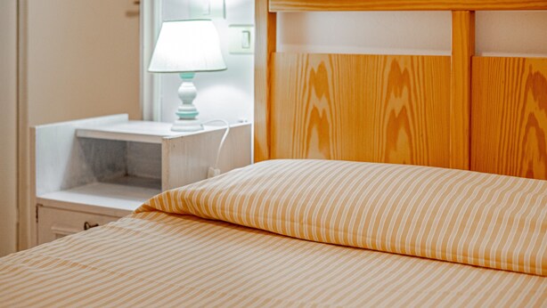 A neatly made bed with striped bedding, a bedside table with a lamp, and a wooden headboard. - ‘La Capannuccia D’ Apartment with Shared Garden, Wi-Fi, and Air Conditioning (Ansedonia)