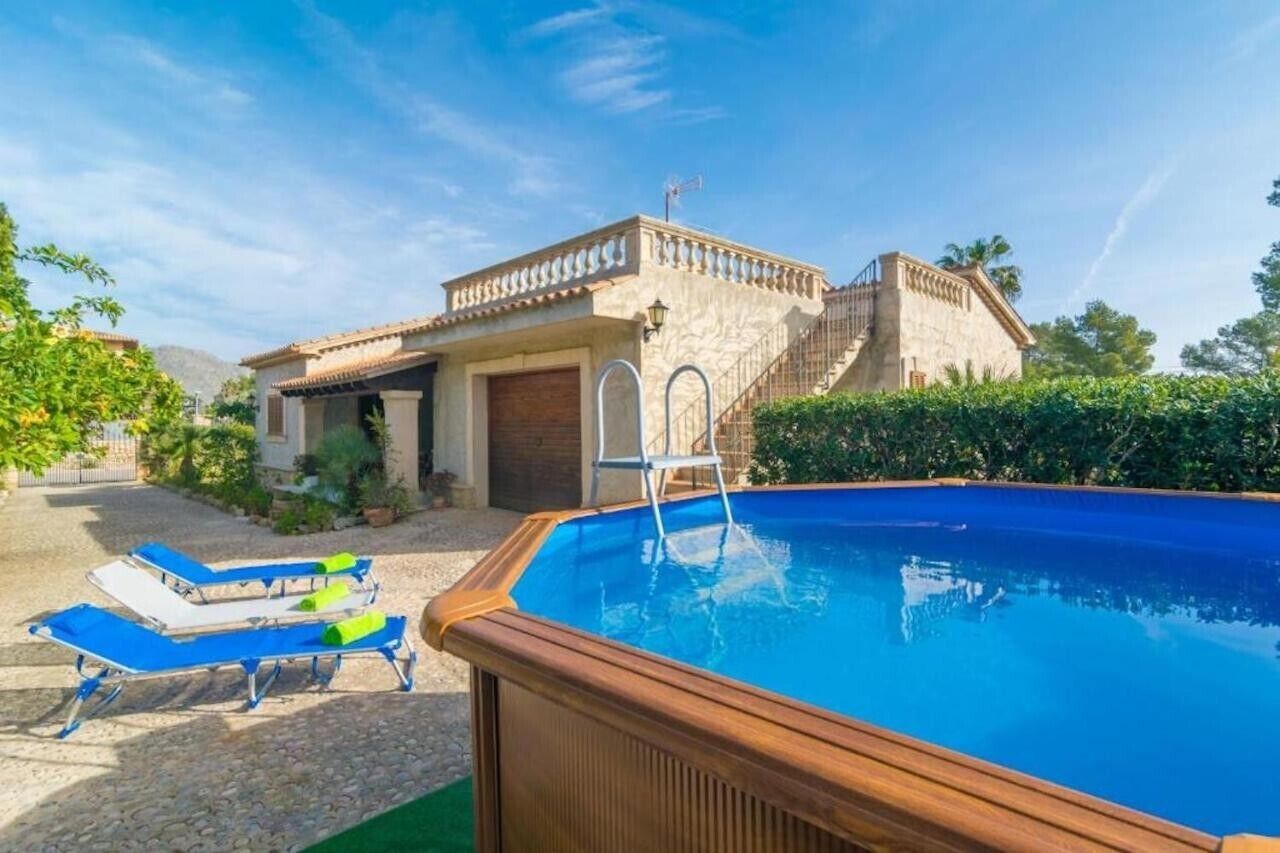 Una piscina con sillones, una terraza de madera, un edificio de piedra con una escalera y un cielo despejado.