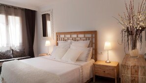 A neatly made bed with white linens, two bedside lamps, a wooden nightstand, a mirror, and a vase with branches on the right side.