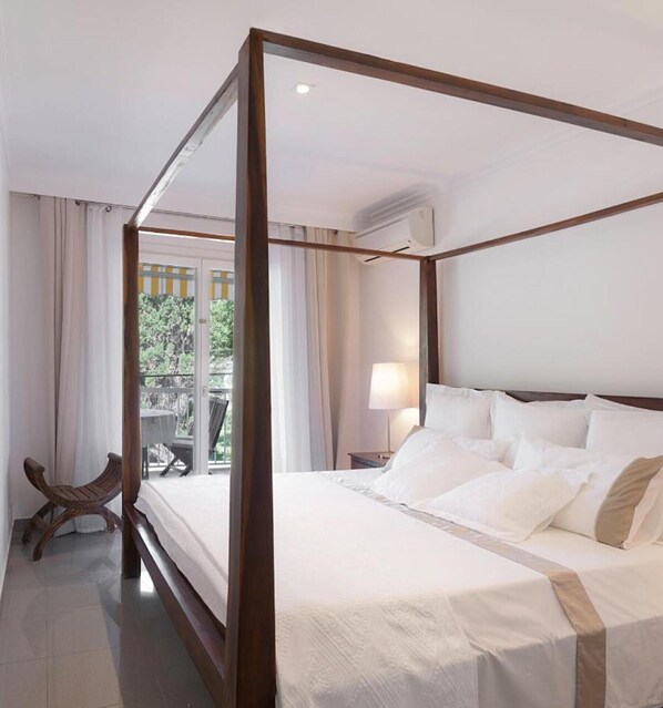 A four-poster bed with white linens, a nightstand with a lamp, and a view of a balcony with outdoor furniture.