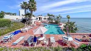 Zona de piscina en un resort con vista al océano, rodeada de palmeras y sillones de descanso.