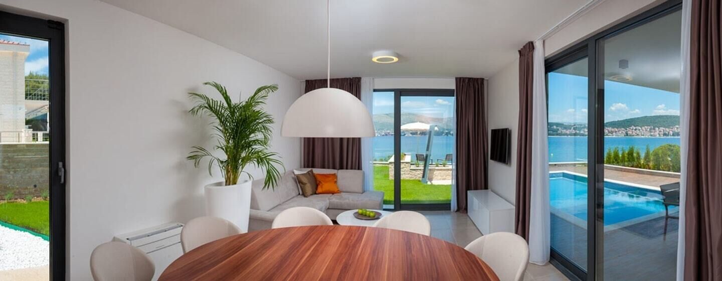 Ein modernes Wohnzimmer mit einem Holzesstisch, weißen Sofas, einer großen Topfpflanze und Blick auf einen Swimmingpool sowie Stadtansicht durch große Glaswände.
