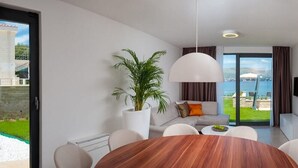 A modern living room with a wooden dining table, white sofas, a large potted plant, and a view of a swimming pool and cityscape through large glass doors. - Villa Safranka - Private villa with pool (Okrug Gornji)