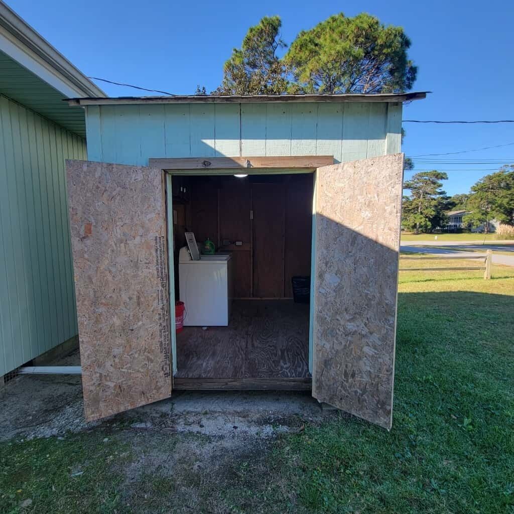 Un petit abri ouvert sur les côtés, avec un intérieur en bois, un lave-linge blanc et une porte donnant sur une cour herbeuse.