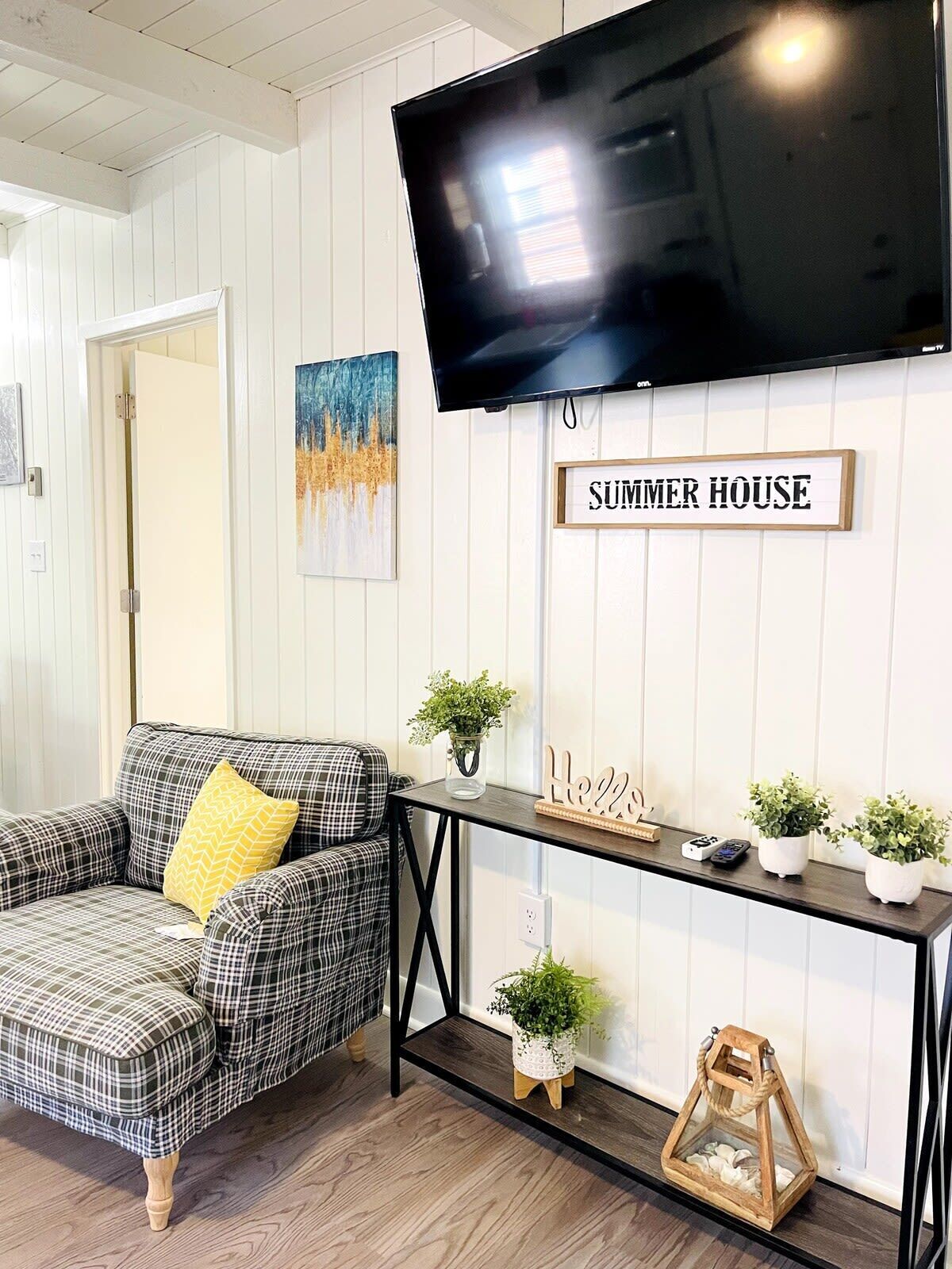 Un salon avec un canapé à carreaux, un téléviseur fixé au mur, une enseigne indiquant « SUMMER HOUSE » et une console sur laquelle sont disposées des plantes et des objets de décoration.