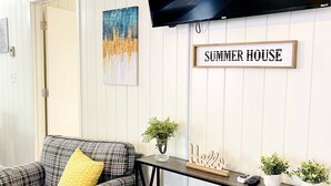 A living room with a plaid sofa, a mounted TV, a 'SUMMER HOUSE' sign, and a console table with plants and decor.