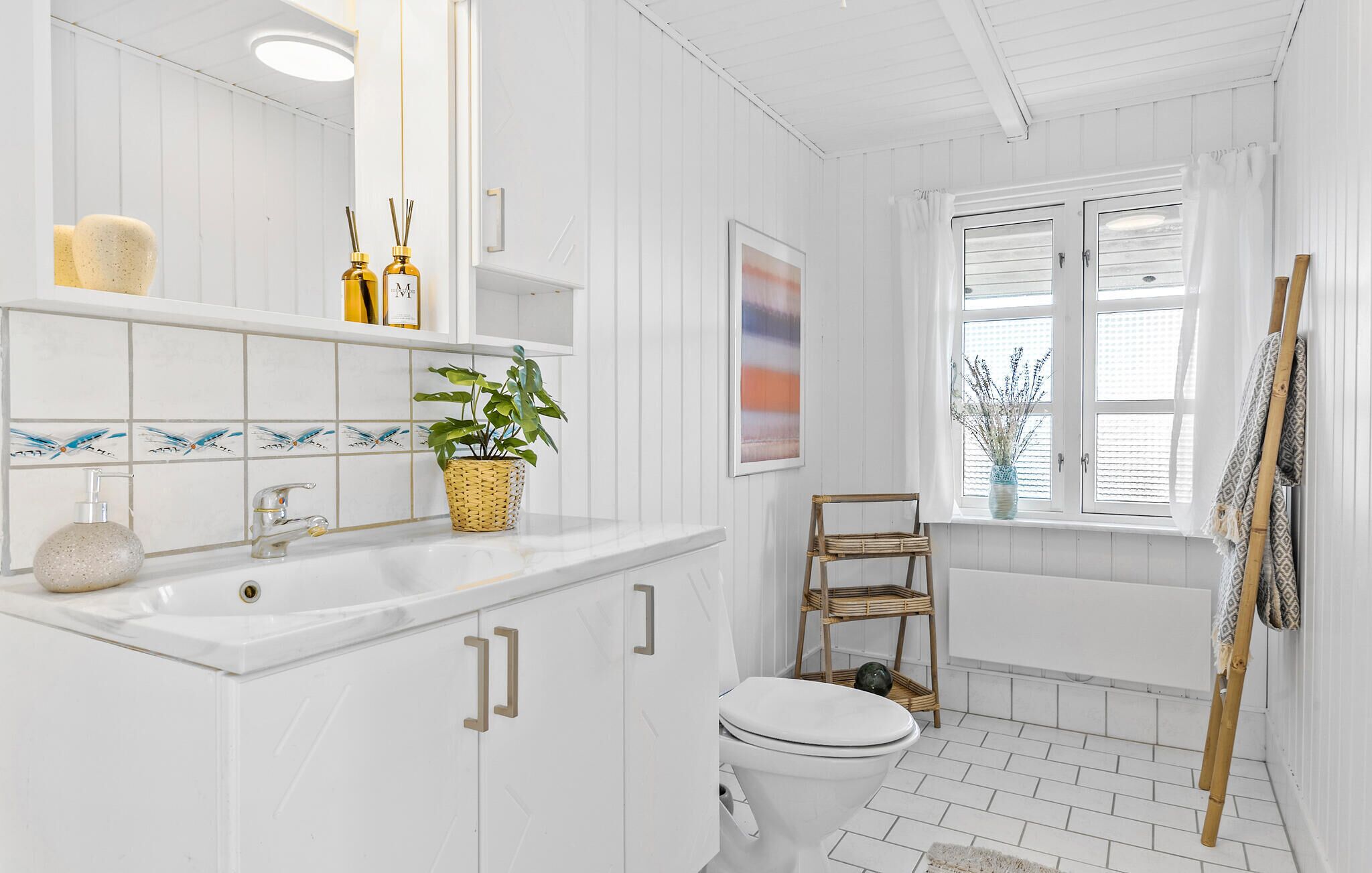 A bathroom with a white sink, mirror, and toilet, a window with white shutters, and a wicker shelf with a plant.