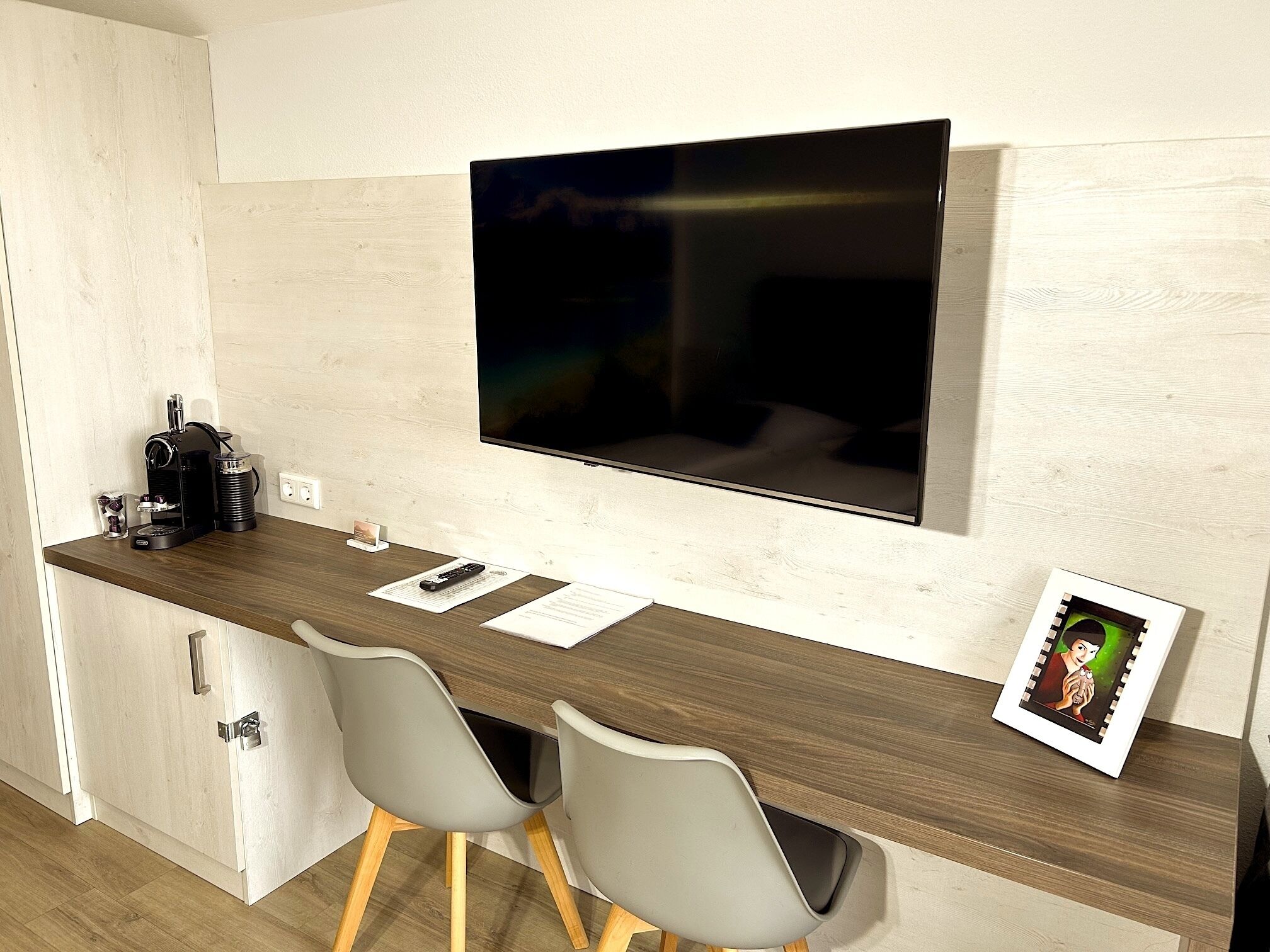A modern kitchen with a wooden countertop, two grey chairs, a TV mounted on the wall, and a small coffee machine.