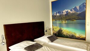 Habitación de hotel con cama, mesita de noche y un amplio ventanal con vistas a una montaña y un lago.