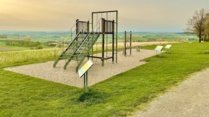 Un parque infantil con estructuras para escalar y columpios, frente a un paisaje rural con campos y un edificio lejano.