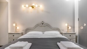 Cama bem arrumada com lençóis brancos e duas toalhas dobradas sobre ela. Há duas mesas de cabeceira com abajures de cada lado da cama.