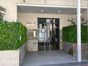 Stella Marina's entrance features a glass door and metal gate, surrounded by lush greenery and neatly trimmed hedges.